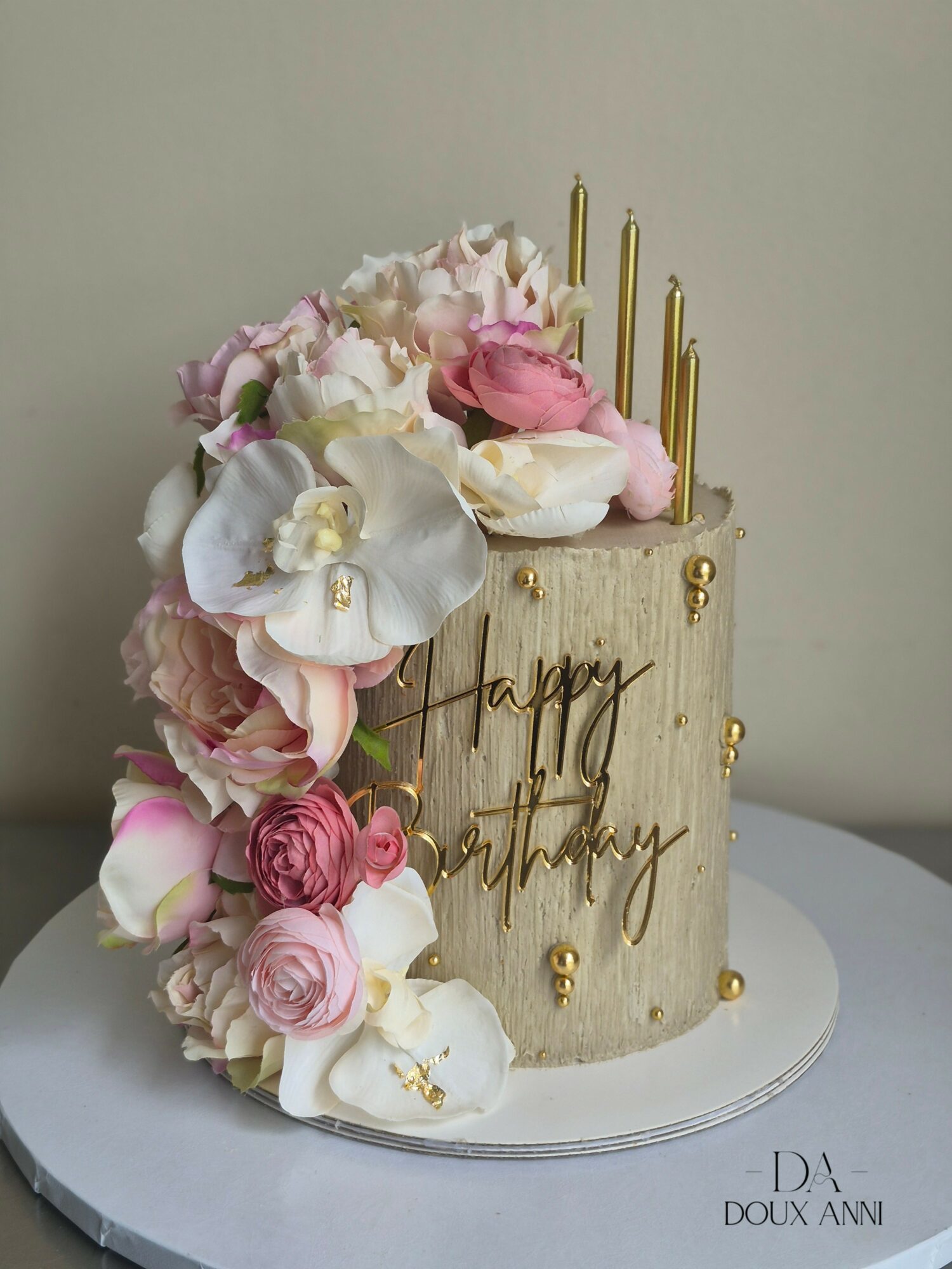 Tall birthday cake decorated with pink and white flowers, gold candles, and a gold 'happy birthday' message, on a white surface.