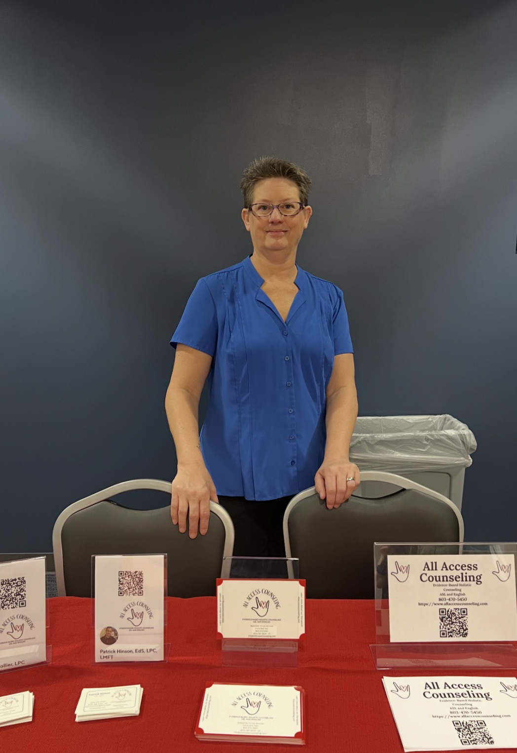 Woman in blue shirt standing behind a table with brochures and a sign, against a dark background.