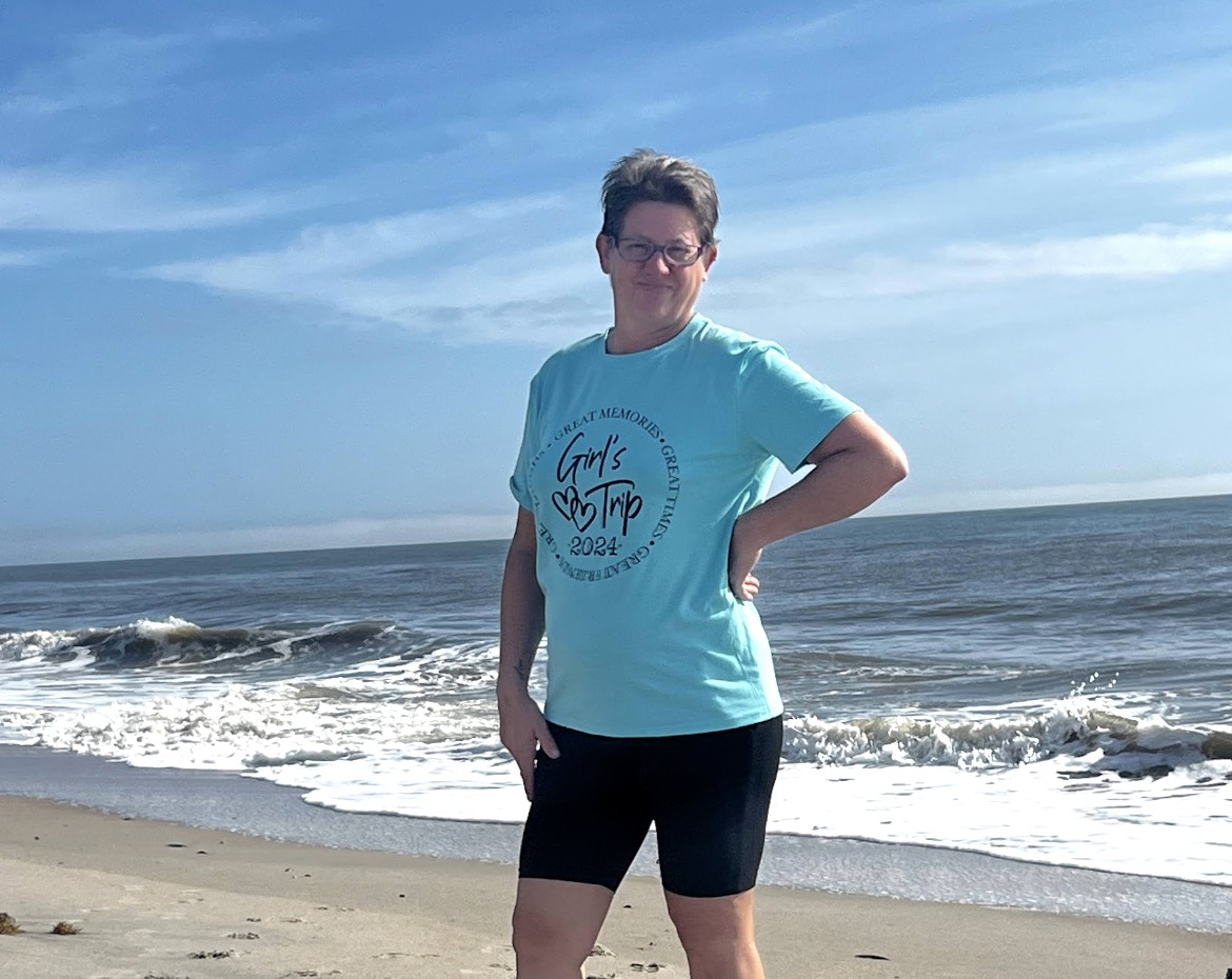 Person standing on a beach with ocean waves and blue sky in the background, wearing a turquoise T-shirt and black shorts.