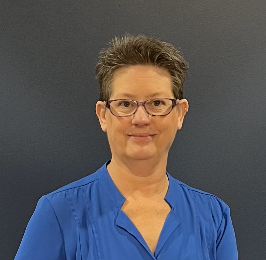 Woman with short hair and glasses wearing a blue top, smiling, against a dark background.