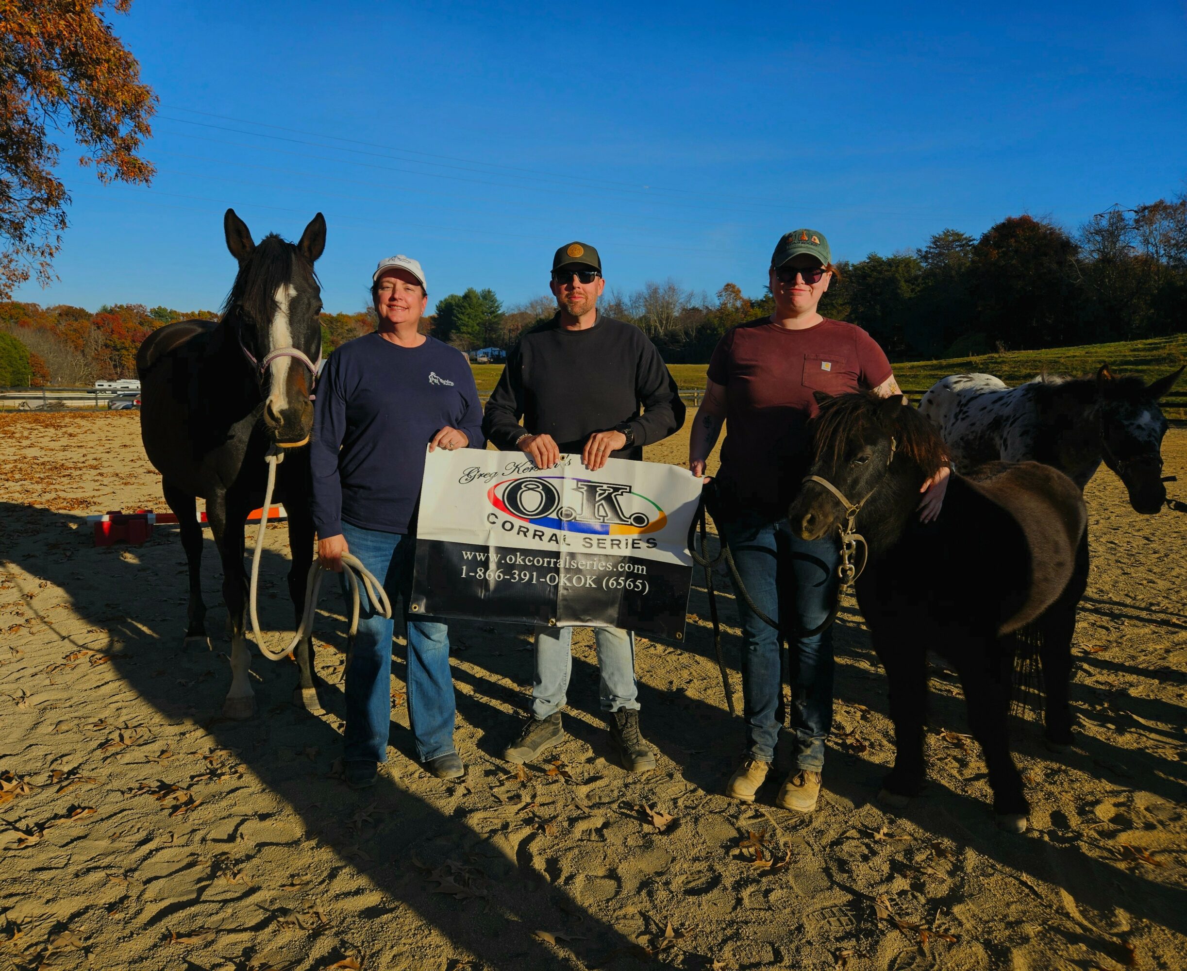 Three people stand with horses outdoors on a dirt field, holding a banner, with trees and a blue sky in background.