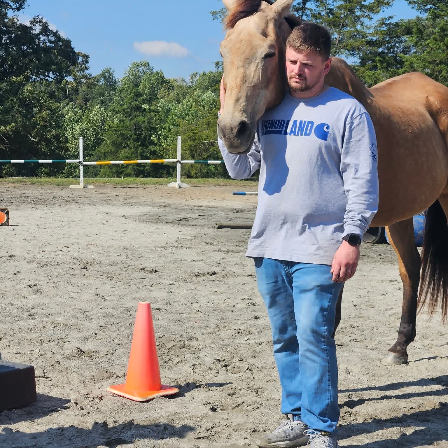 Man standing outdoors with a horse resting its head on his shoulder, orange cone nearby, trees in background.