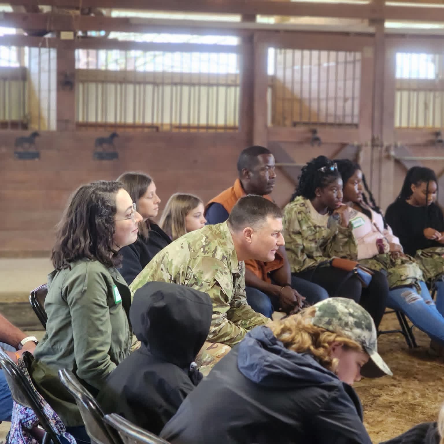Group of diverse people sitting on chairs indoors, some wearing military camouflage, in a rustic setting with wooden walls and windows.