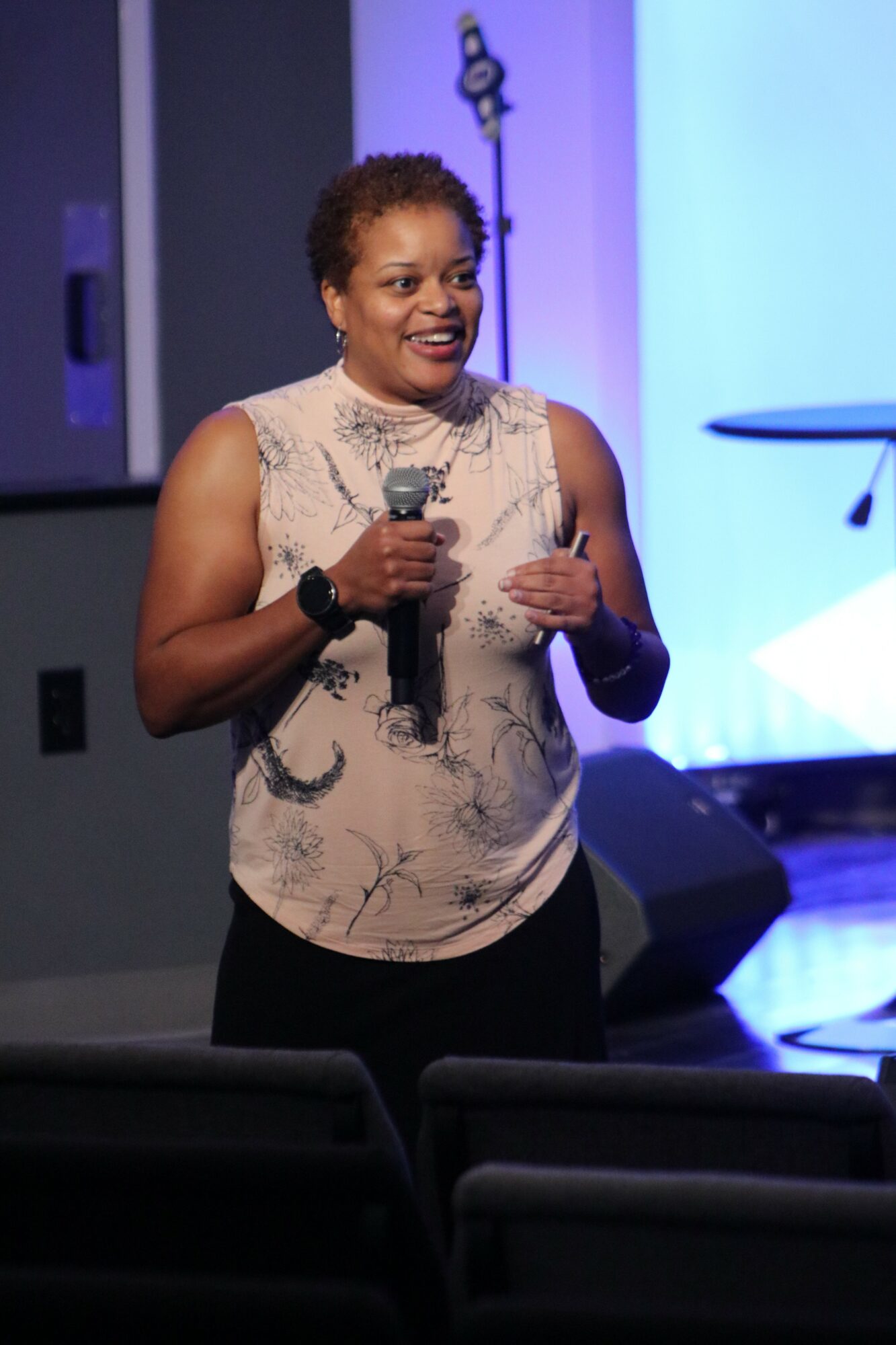 Woman speaking into a microphone on stage, wearing a sleeveless top, with a blue-lit background.