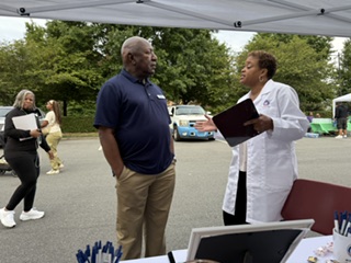 Two people, one in a navy shirt and the other in a white coat, are talking outdoors under a canopy, with cars and trees in background.
