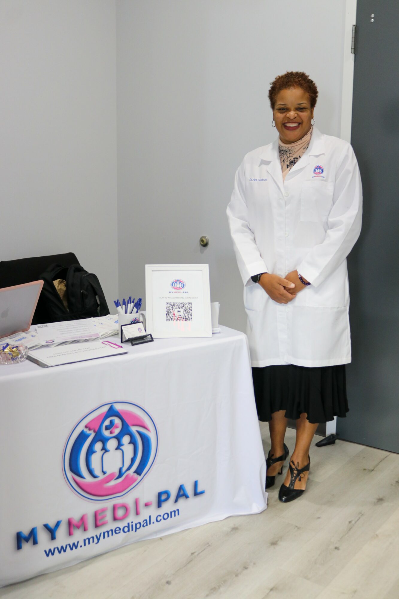 Woman in white lab coat standing next to a table with medical materials, smiling, in a clinical setting.