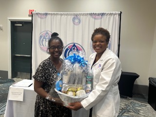 Two women smiling, one in a patterned dress and the other in a white coat, holding a gift basket with a blue bow.