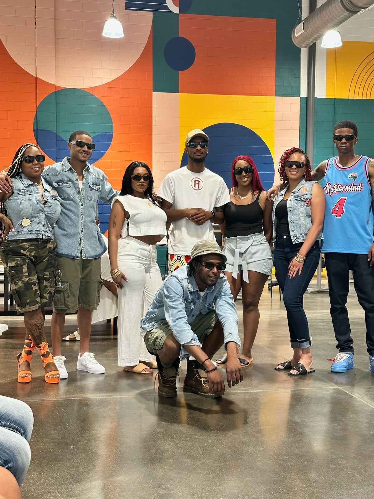 Group of eight people standing indoors in front of colorful geometric wall, some smiling, some looking serious, casual clothing, diverse ages and styles.