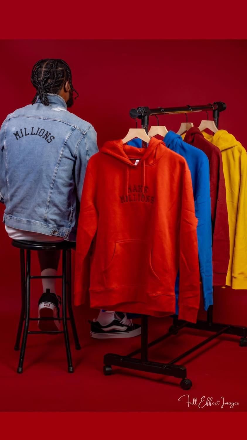 Person with braided hair wearing a denim jacket labeled 'MILLIONS' stands next to colorful hoodies on a rack against a red background.
