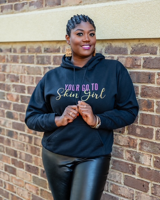 Woman with braided hair wearing a black hoodie and leather pants standing against a brick wall.