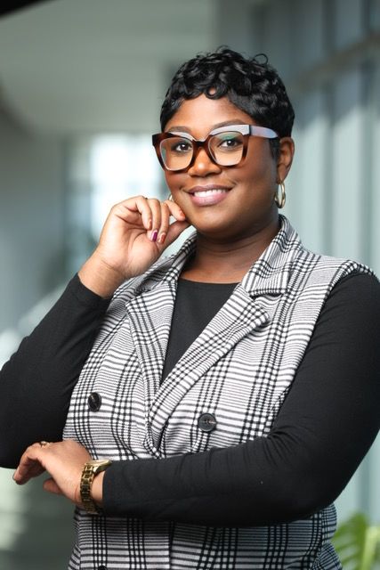 Woman with short curly hair and glasses smiling, wearing a black and white checkered blazer, indoors.