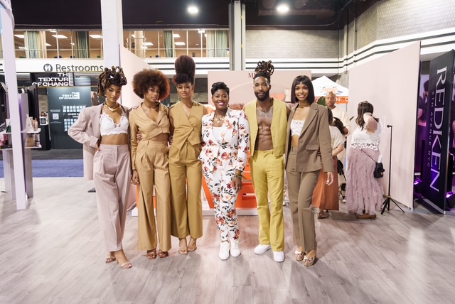 Group of seven women standing together indoors, some wearing stylish outfits, in a well-lit space with signs and displays.