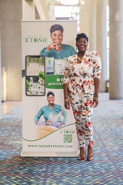 Woman in floral jumpsuit standing next to a promotional display with a woman on it, in a spacious indoor setting.