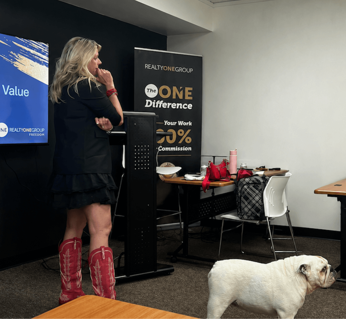Woman with long hair wearing red boots and black dress stands near a presentation screen and banner, with a bulldog nearby.