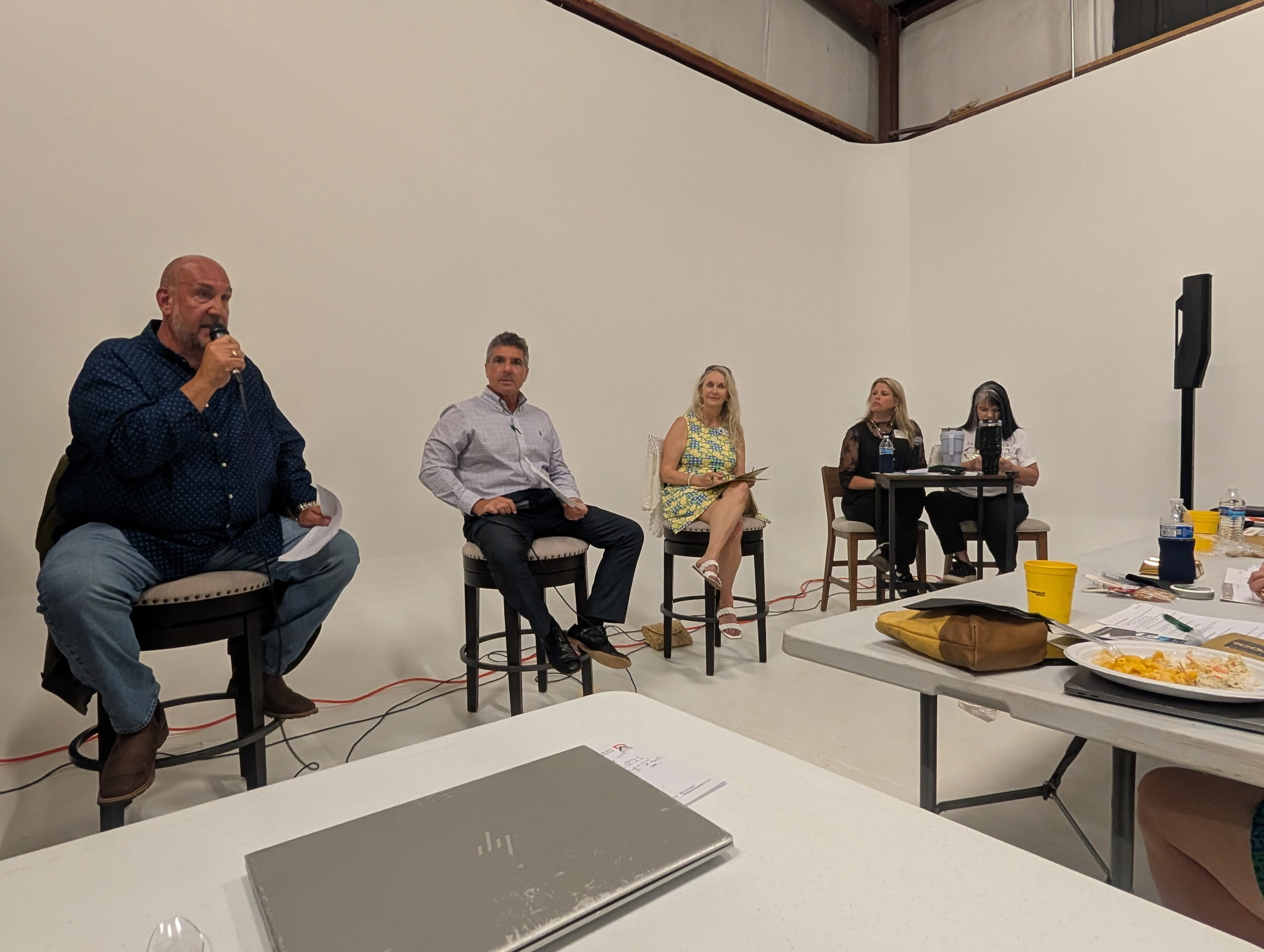 Four people seated in a row, one speaking into a microphone, in a plain white room with tables and food.
