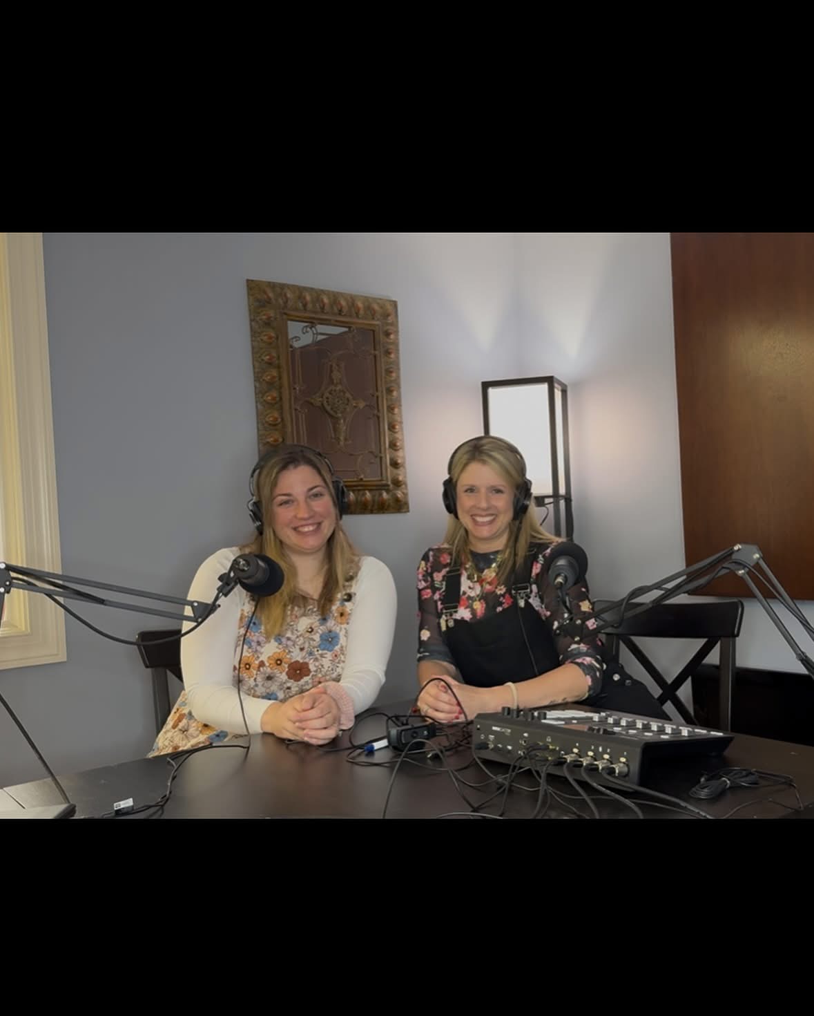 Two women sit at a table with microphones and audio equipment, smiling in a recording studio with a mirror and lamp behind them.
