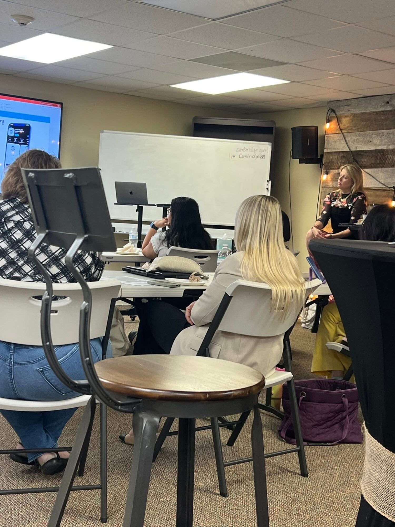 Classroom with students seated at desks, a teacher at the front, whiteboard, and a screen displaying a presentation.