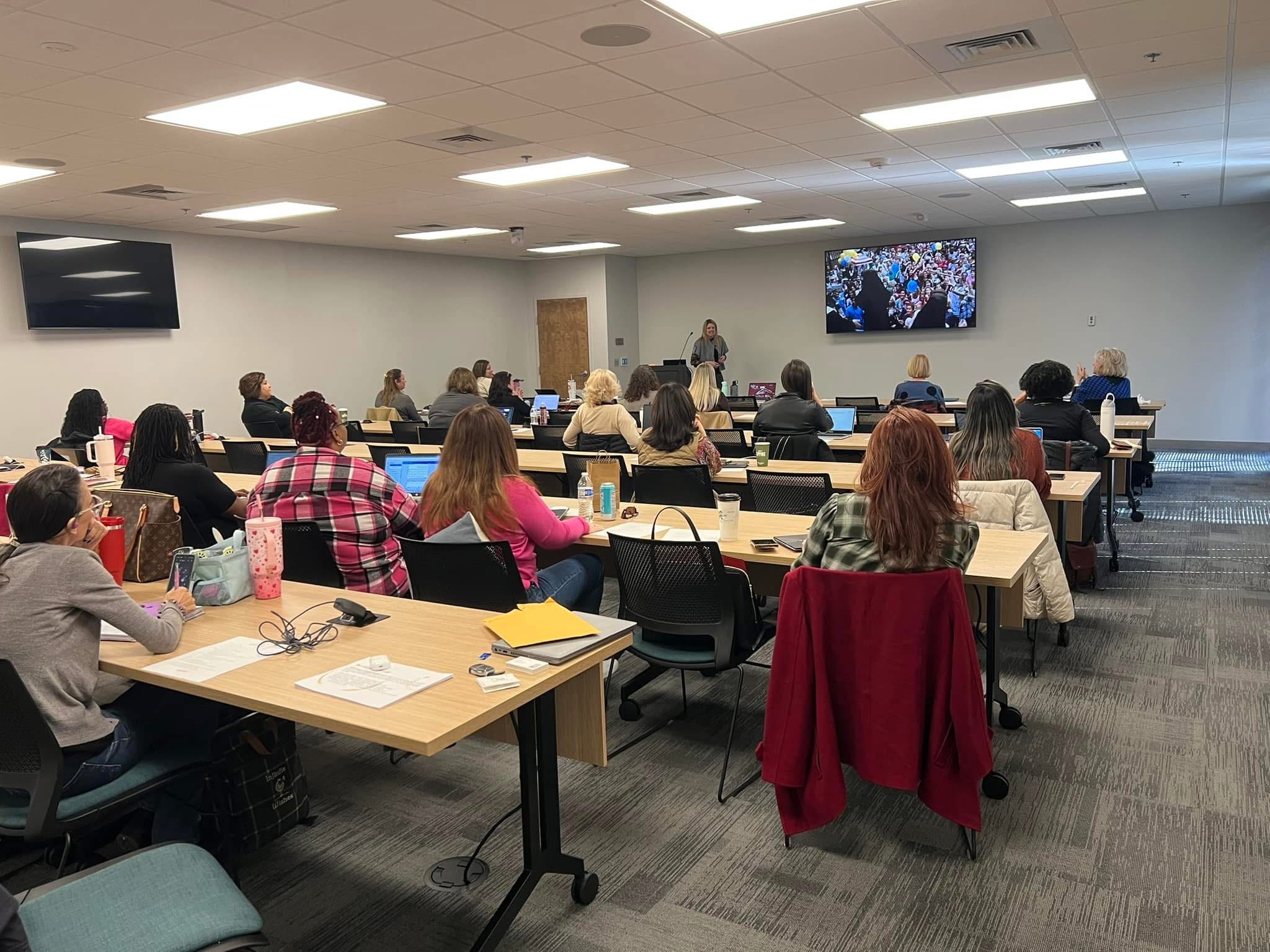 People seated at tables in a conference room watching a presentation on a screen, with some taking notes.
