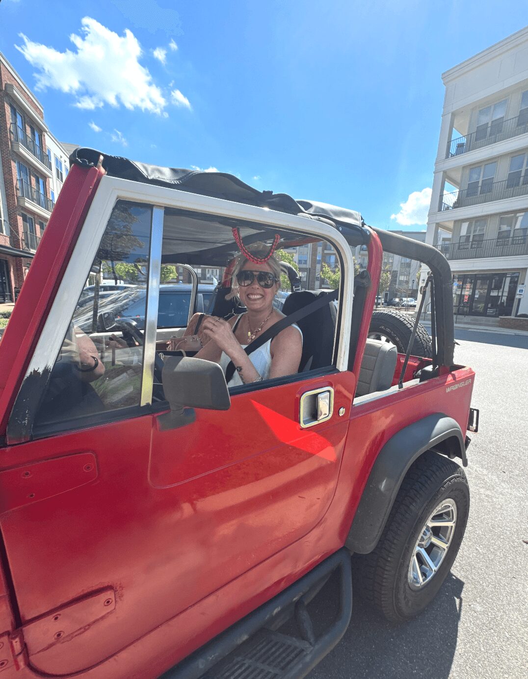 Woman sitting in a red convertible car, smiling, with buildings and blue sky in the background.