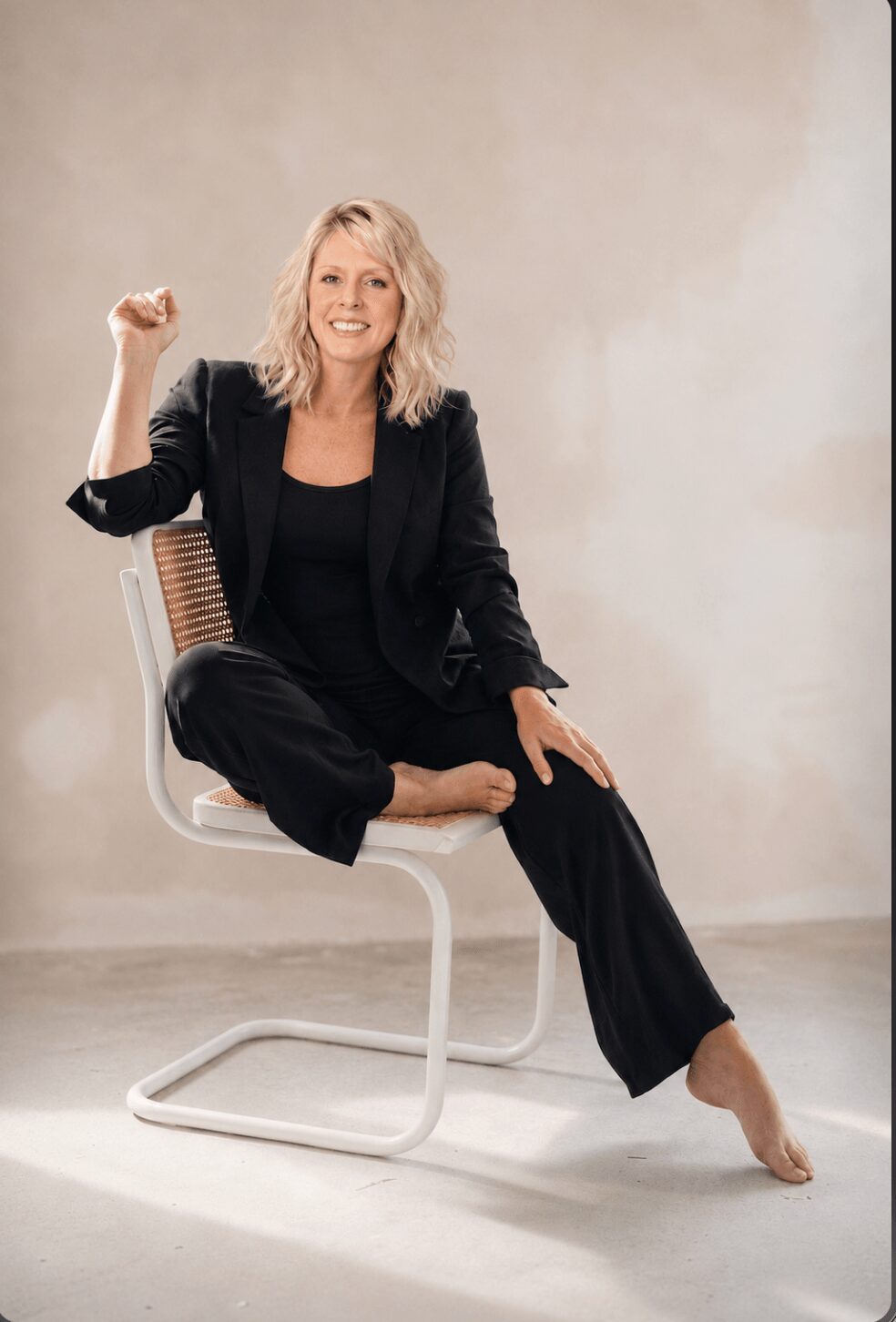 Woman sitting on a chair, smiling, with one arm raised and fist clenched, wearing a black suit, barefoot, against a plain background.