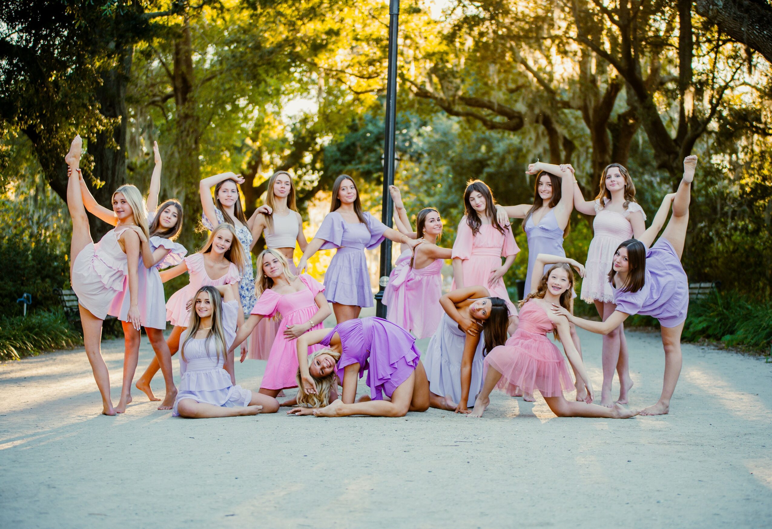 Group of young girls in colorful dresses posing outdoors in a park with trees and sunlight, some standing and some sitting or kneeling.