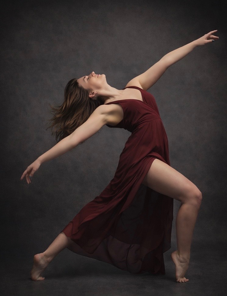 A woman in a flowing burgundy dress dances with arms extended and head tilted back against a dark background.