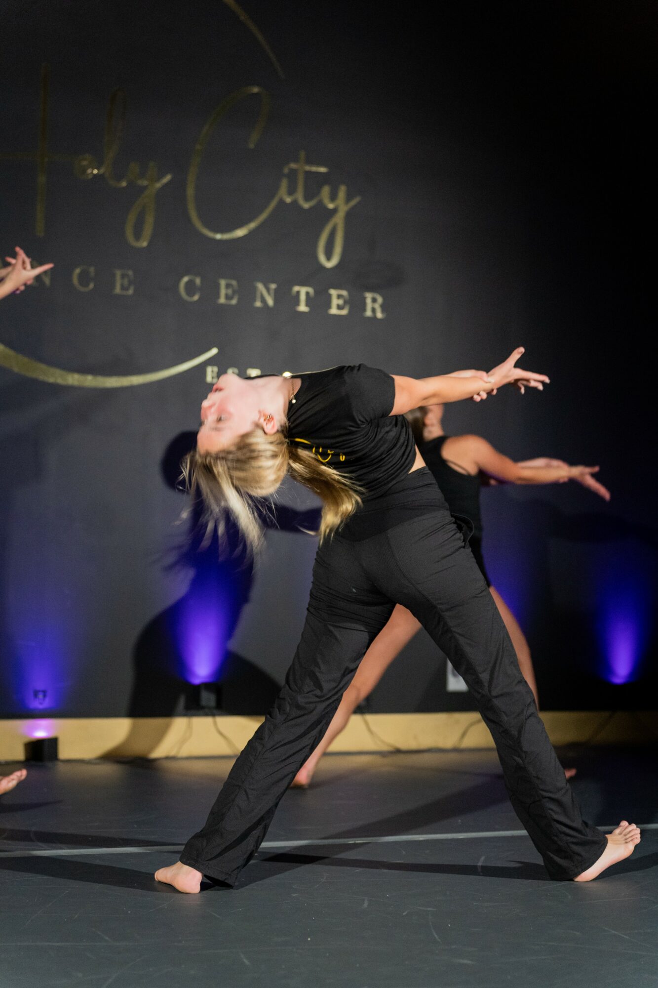 Two dancers perform on stage, one leaning back with arms extended, the other partially visible behind, with a dark background and stage lighting.