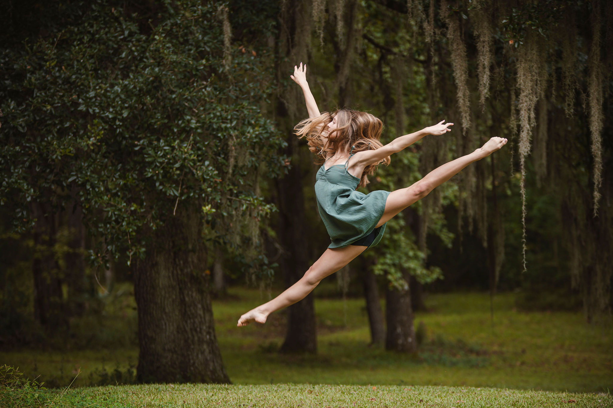 Girl jumping in a park with trees and moss hanging from branches, arms and legs extended mid-air.