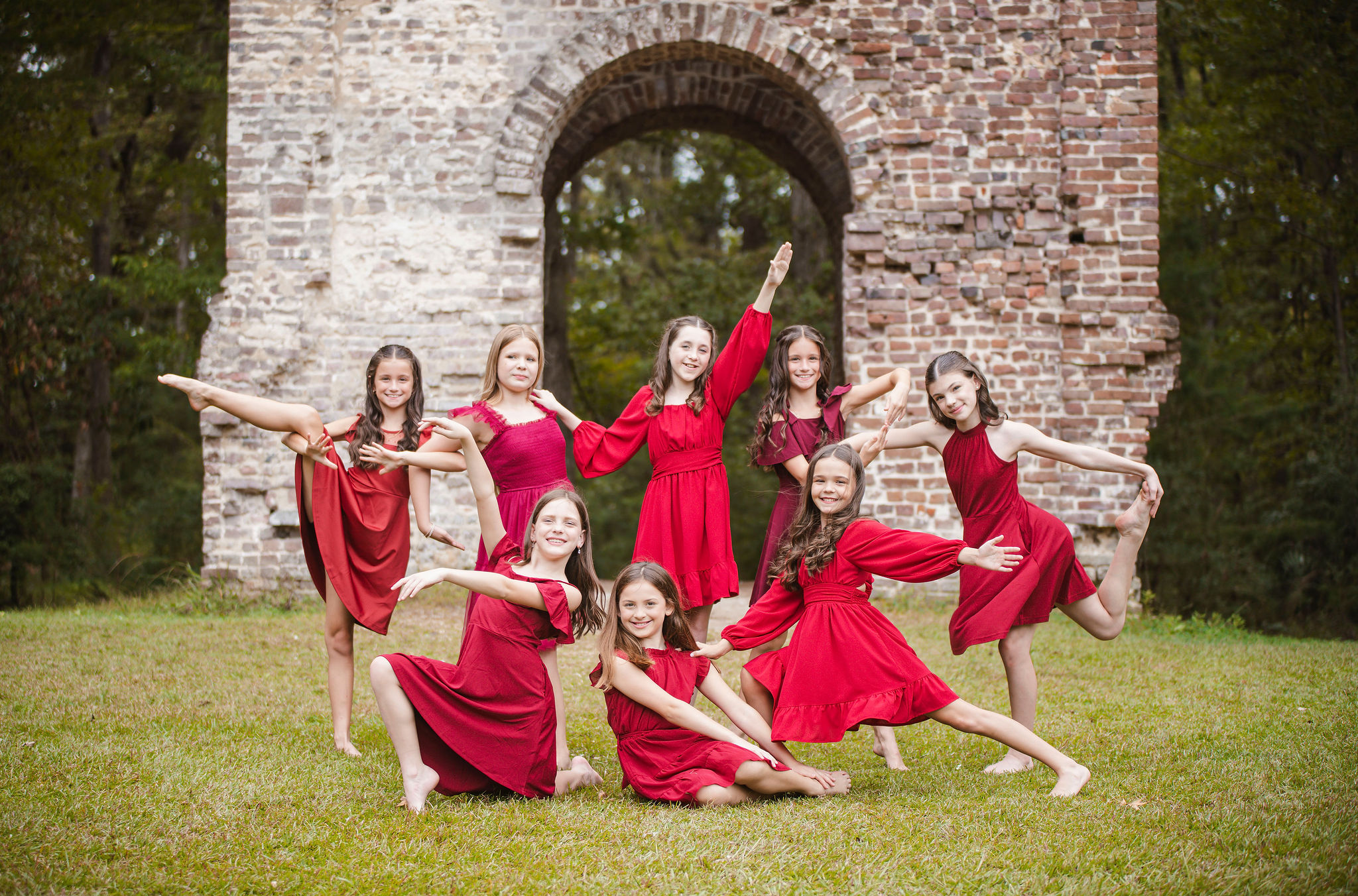 Group of young girls in red dresses performing dance poses outdoors in front of a brick archway.