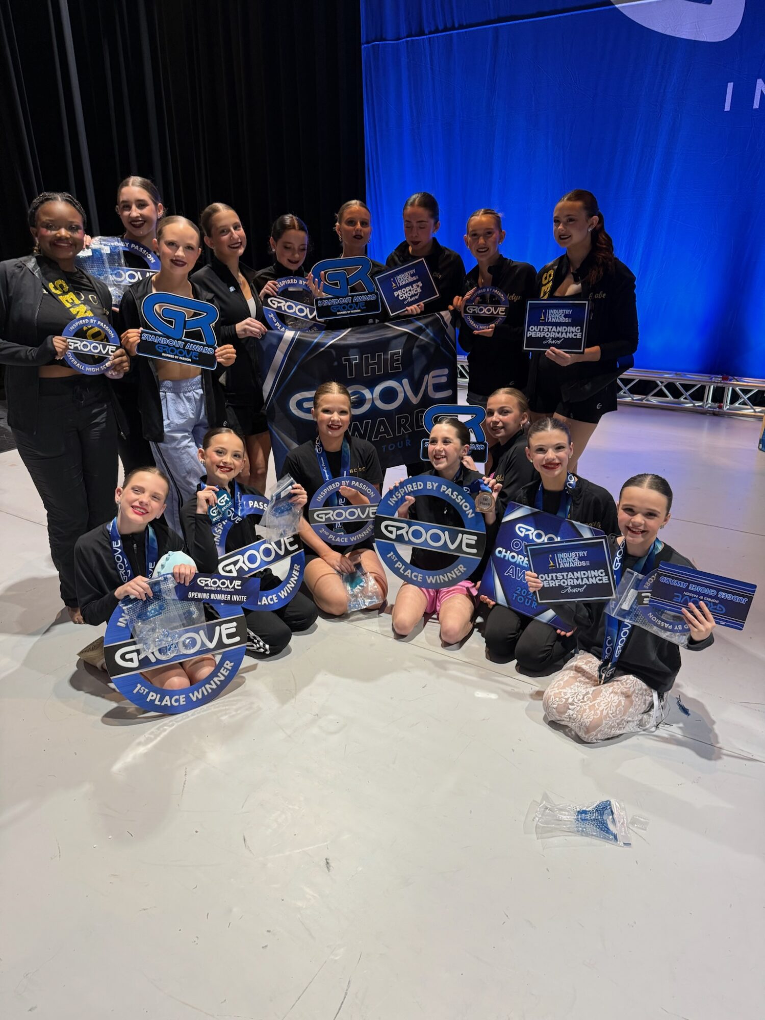 Group of young dancers holding awards and certificates on stage, smiling, with a blue backdrop and stage lighting.