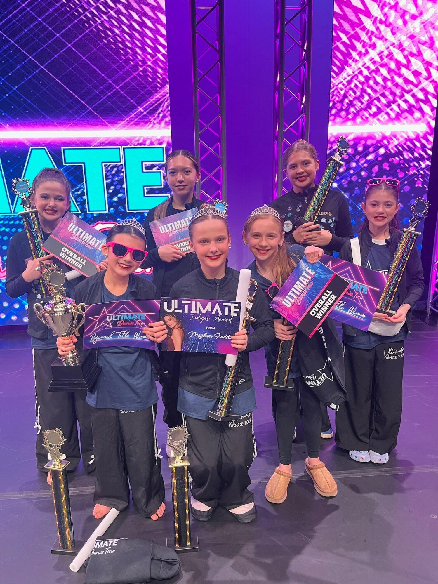 Group of seven young girls holding trophies and certificates on stage with a vibrant purple and blue background.