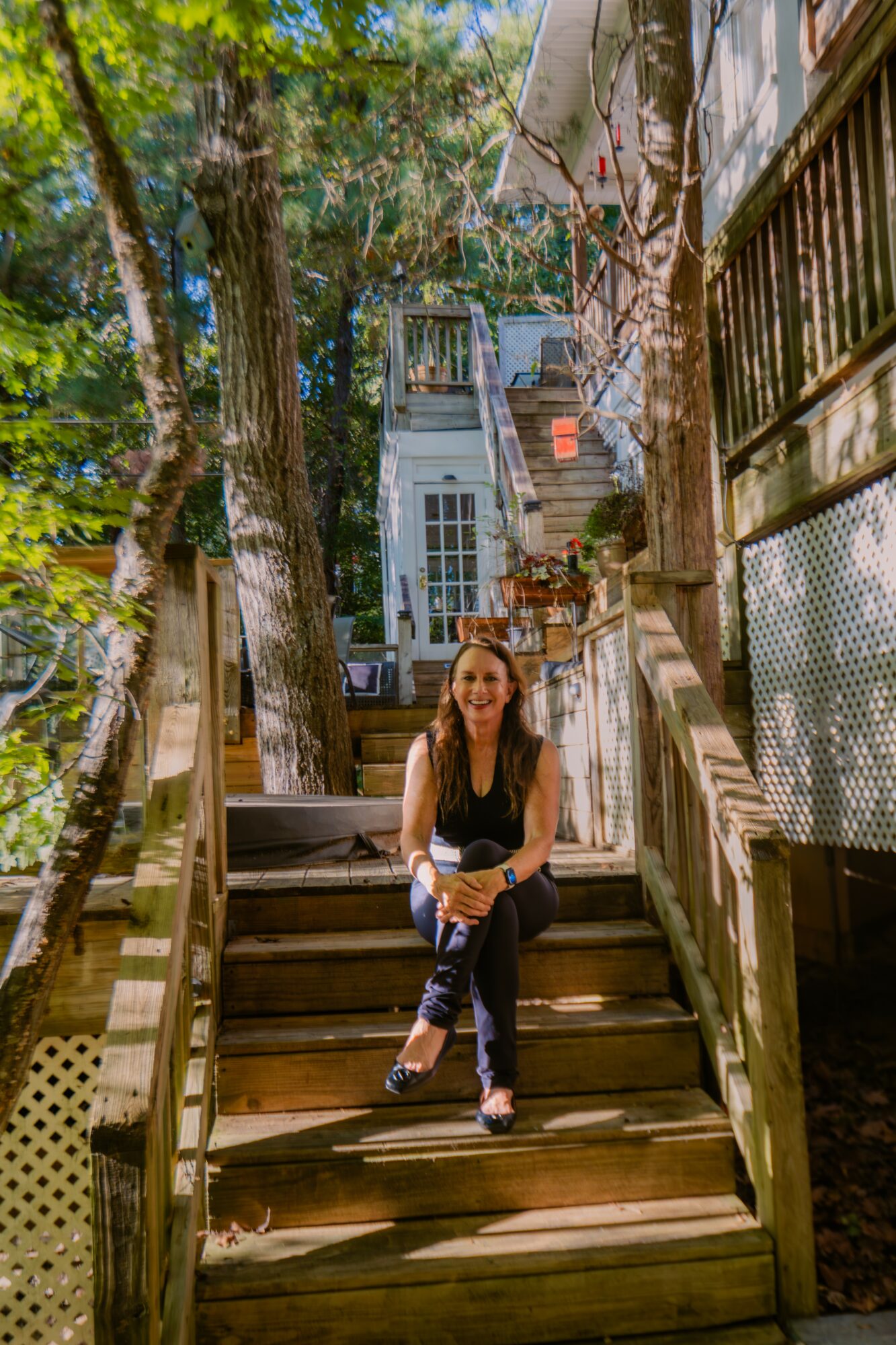 Woman sitting on wooden stairs outdoors, trees and house in background, sunlight filtering through leaves.