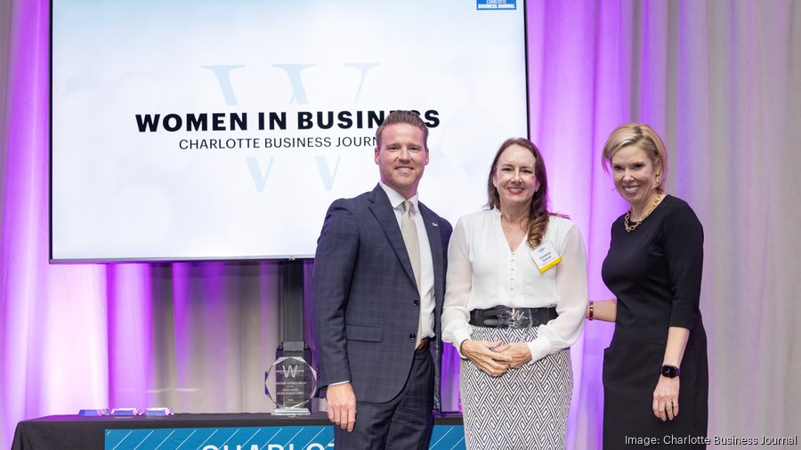 Three people standing in front of a screen that reads 'Women in Business' at a conference, smiling.