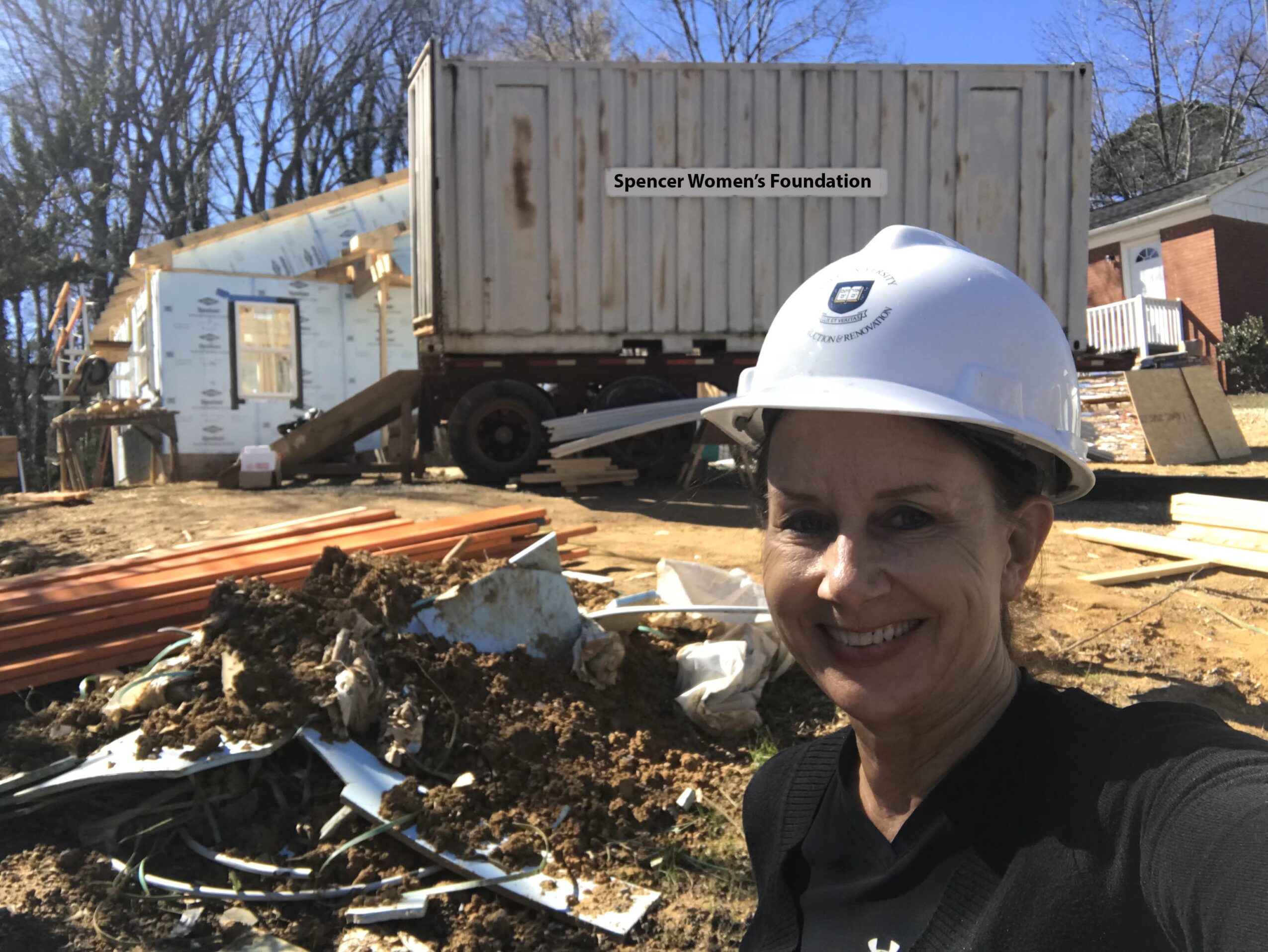 Woman wearing a white safety helmet smiling outdoors with construction debris and a container in the background.