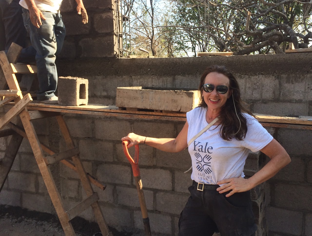 Woman smiling with sunglasses holding a shovel at a construction site, standing in front of a brick wall.