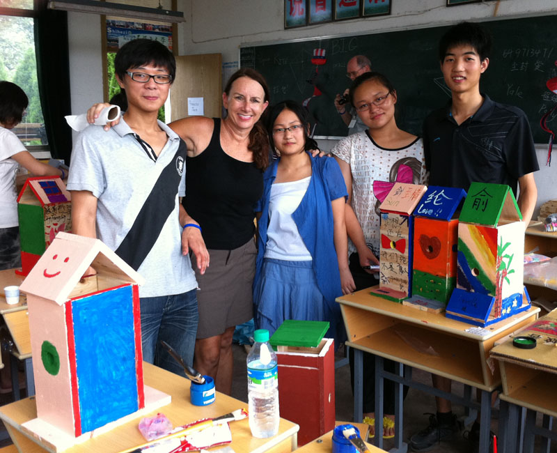 Group of five people, including students and a teacher, standing in a classroom with colorful projects on tables.