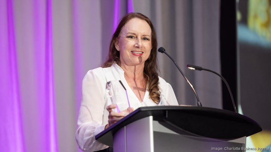 Woman speaking at a podium with a microphone, smiling, in front of a purple curtain.