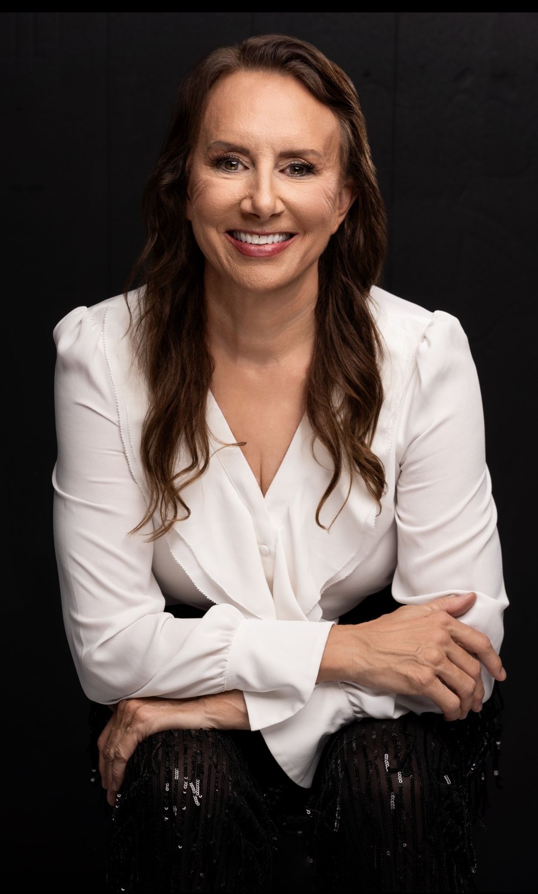 Woman with long wavy hair smiling, wearing a white blouse, sitting with arms crossed against a dark background.