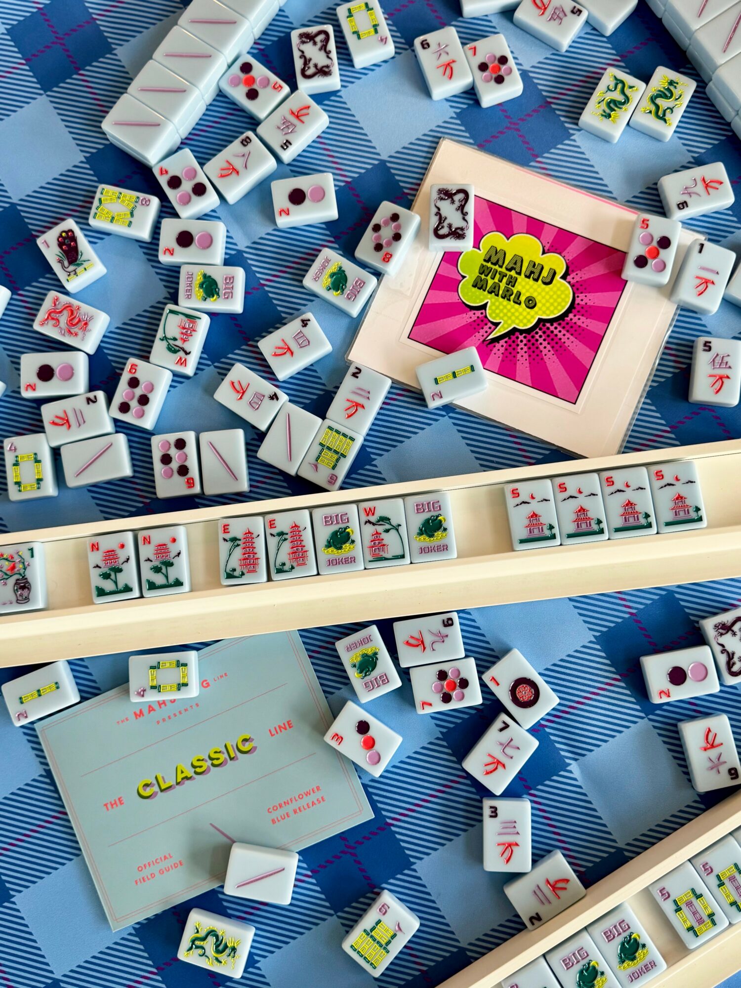 Mahjong tiles scattered on a blue checkered surface with a pink card reading 'Mahjong' and a row of tiles at the bottom.