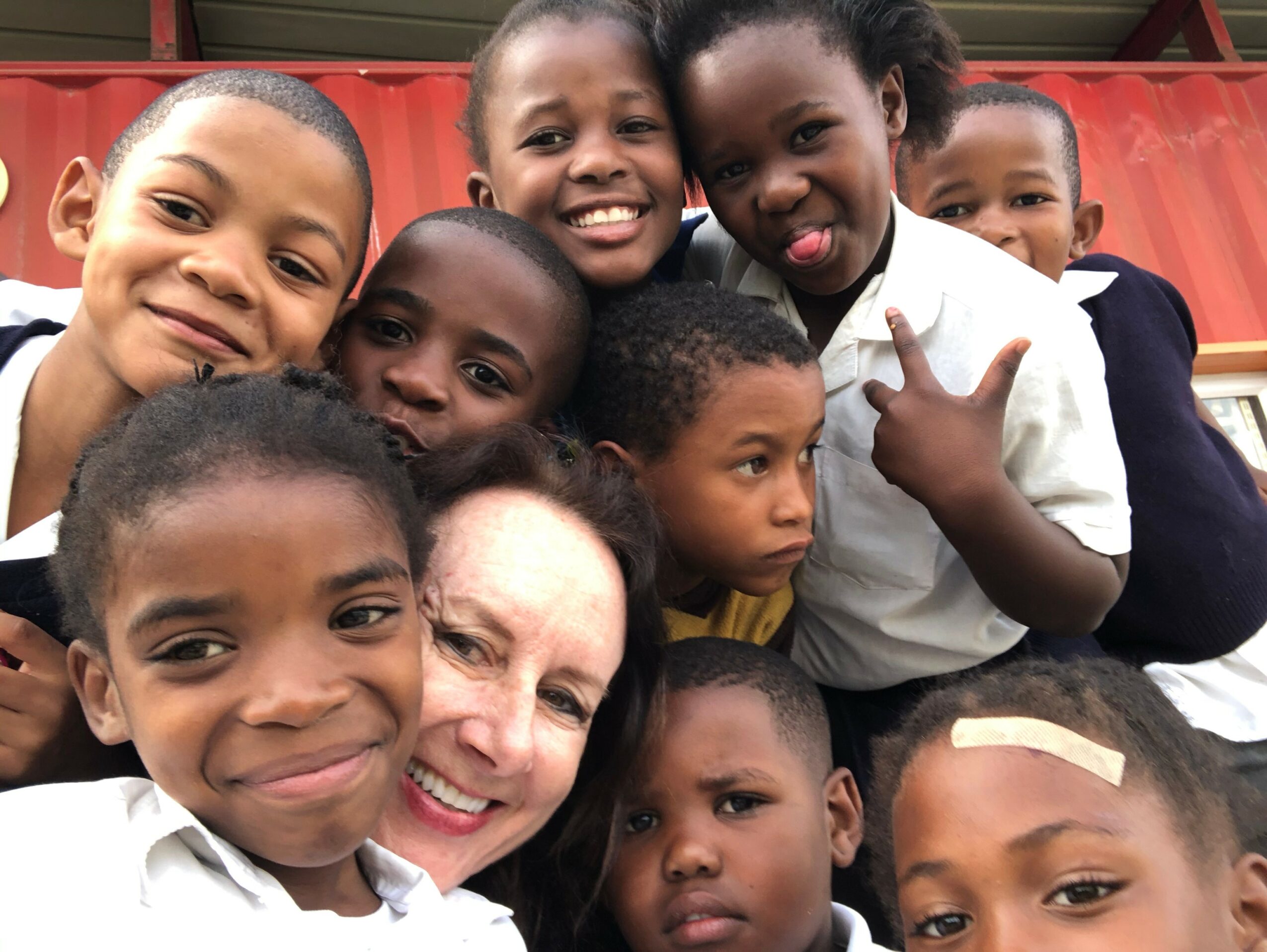 Group of children and a woman smiling and posing together outdoors, some making peace signs.