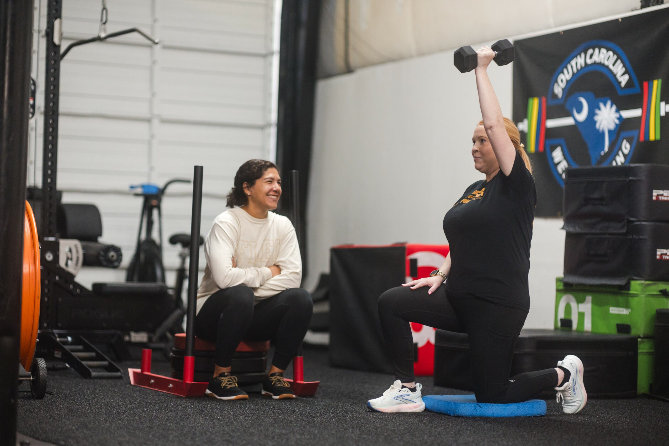 Two women exercising indoors; one is sitting on a red step platform, the other is kneeling and lifting a dumbbell overhead.