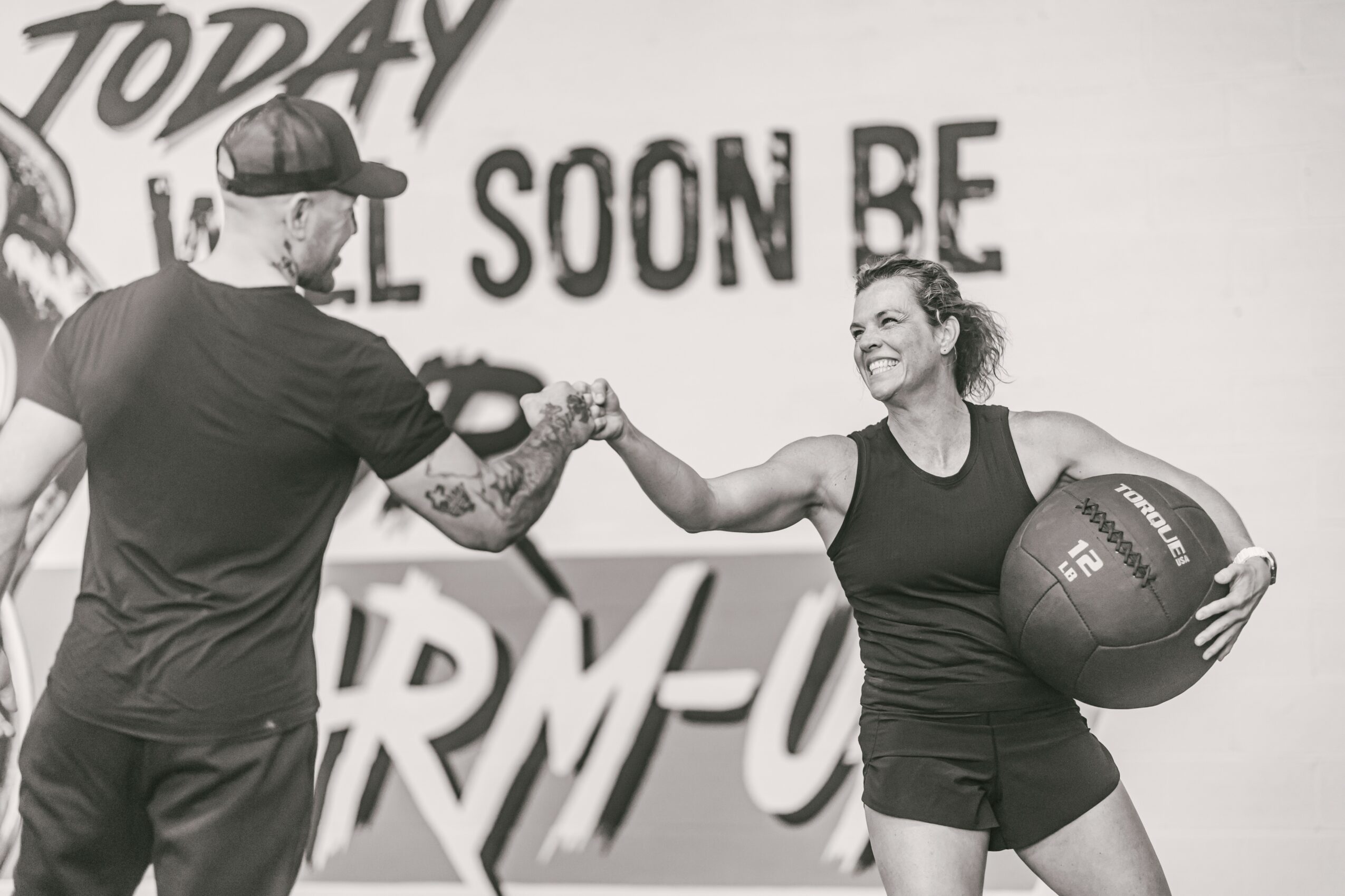 Two people shake hands in a gym, one holding a medicine ball, smiling, with a motivational wall in the background.
