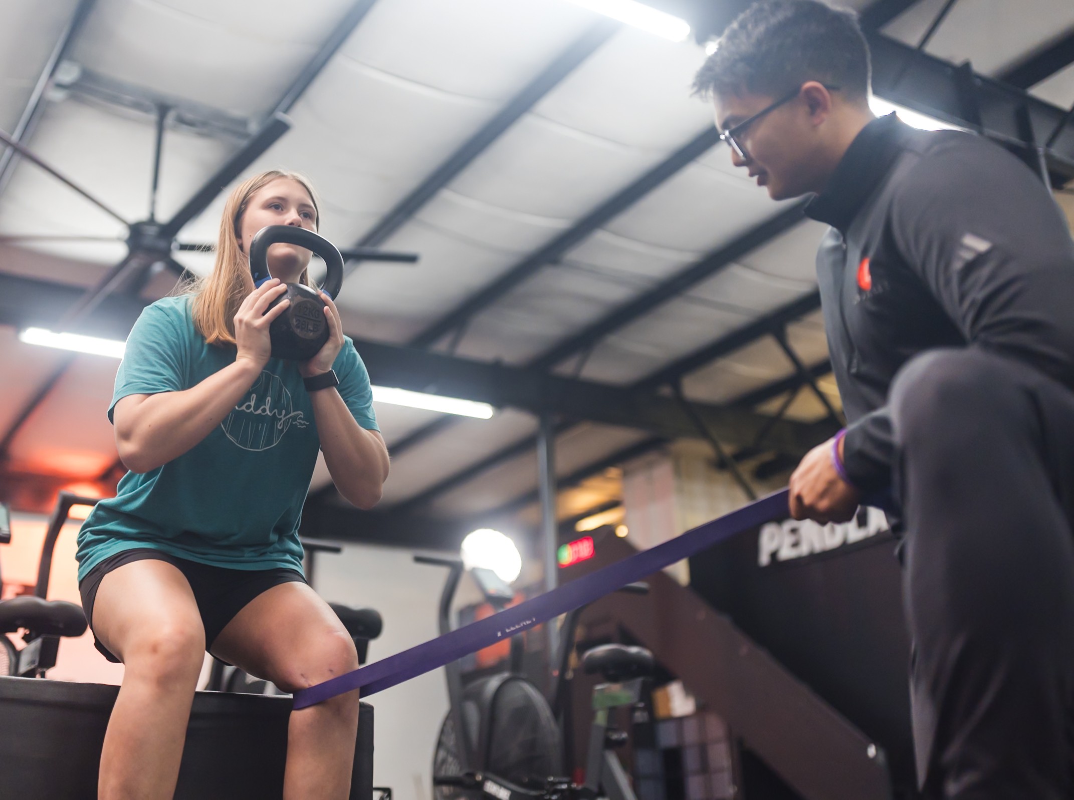 A woman holding a camera crouches on a gym floor, facing a man in workout attire, with gym equipment in background.