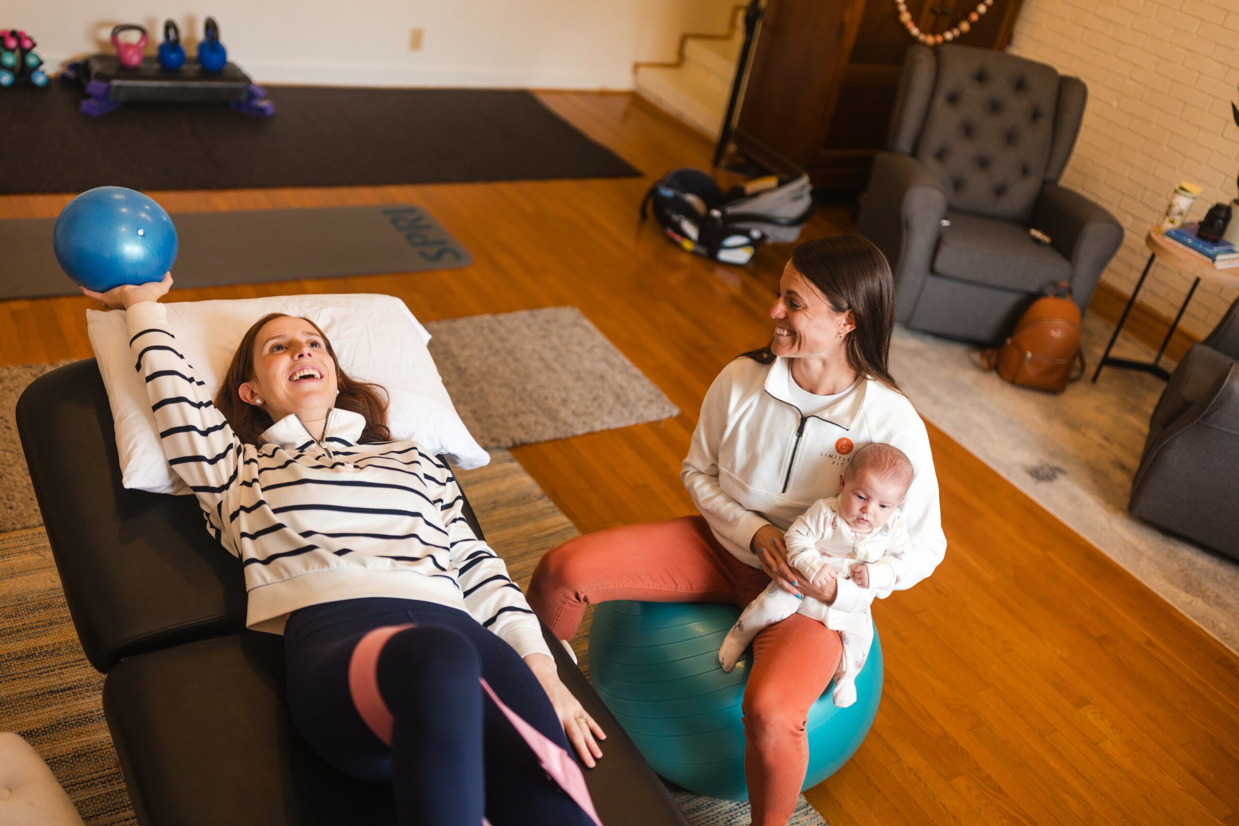 Two women and a baby in a living room with wooden floors, one woman lying on a black couch, the other sitting on an exercise ball holding the baby.