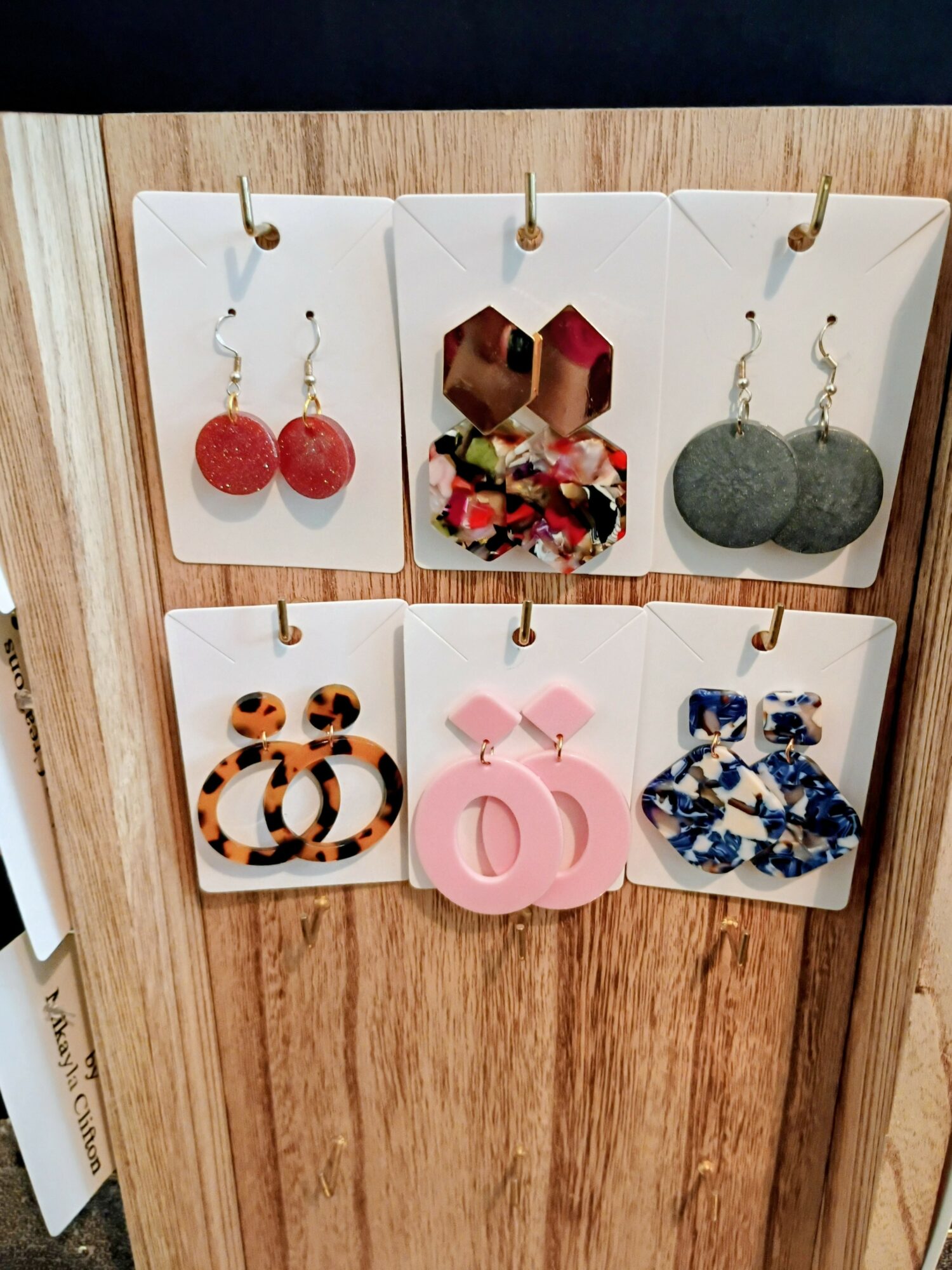 Six pairs of earrings displayed on white cards attached to a wooden board, featuring various shapes and colors.