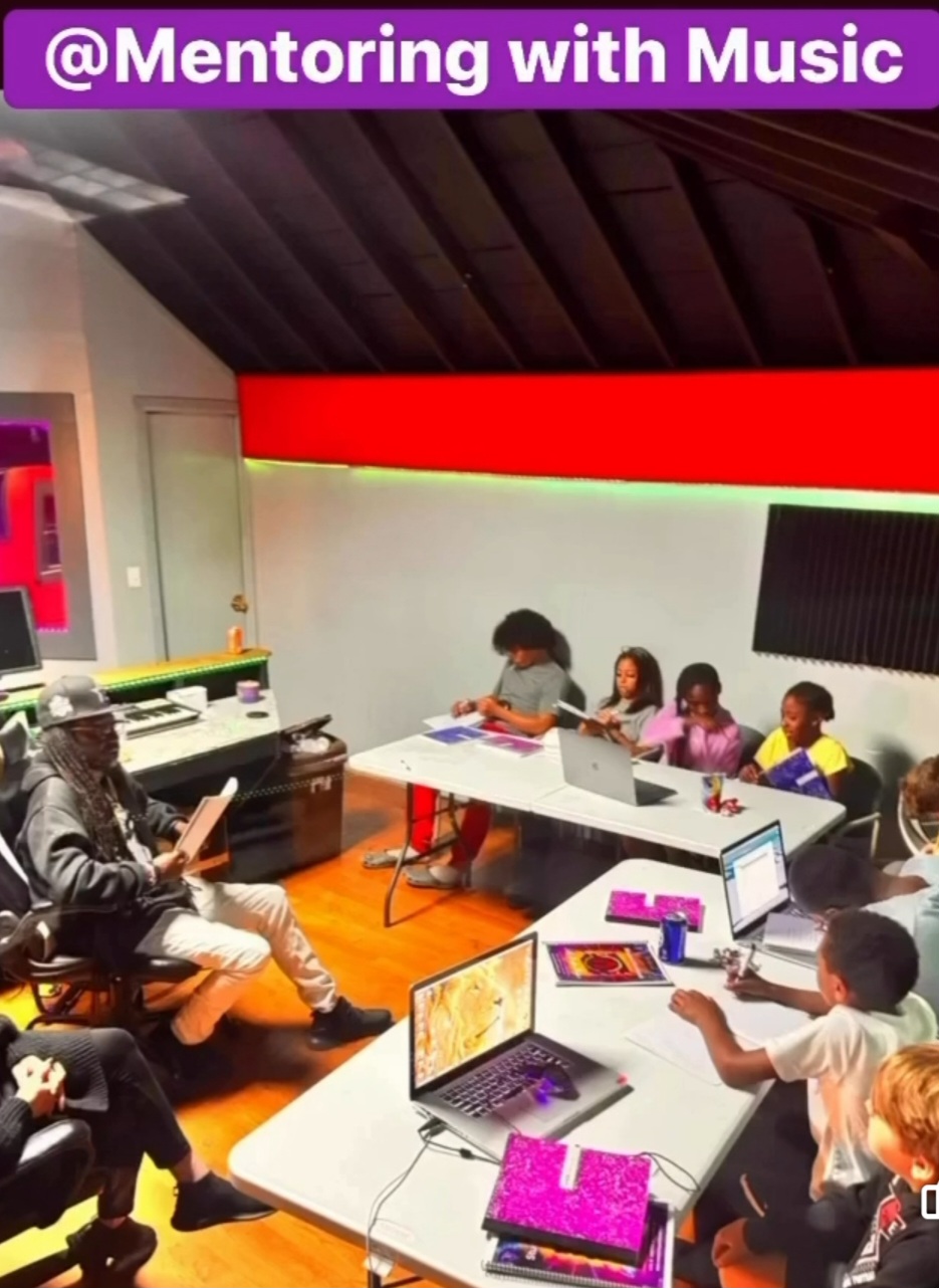 Group of children and an adult in a music mentoring session with laptops and notebooks in a room with colorful walls.