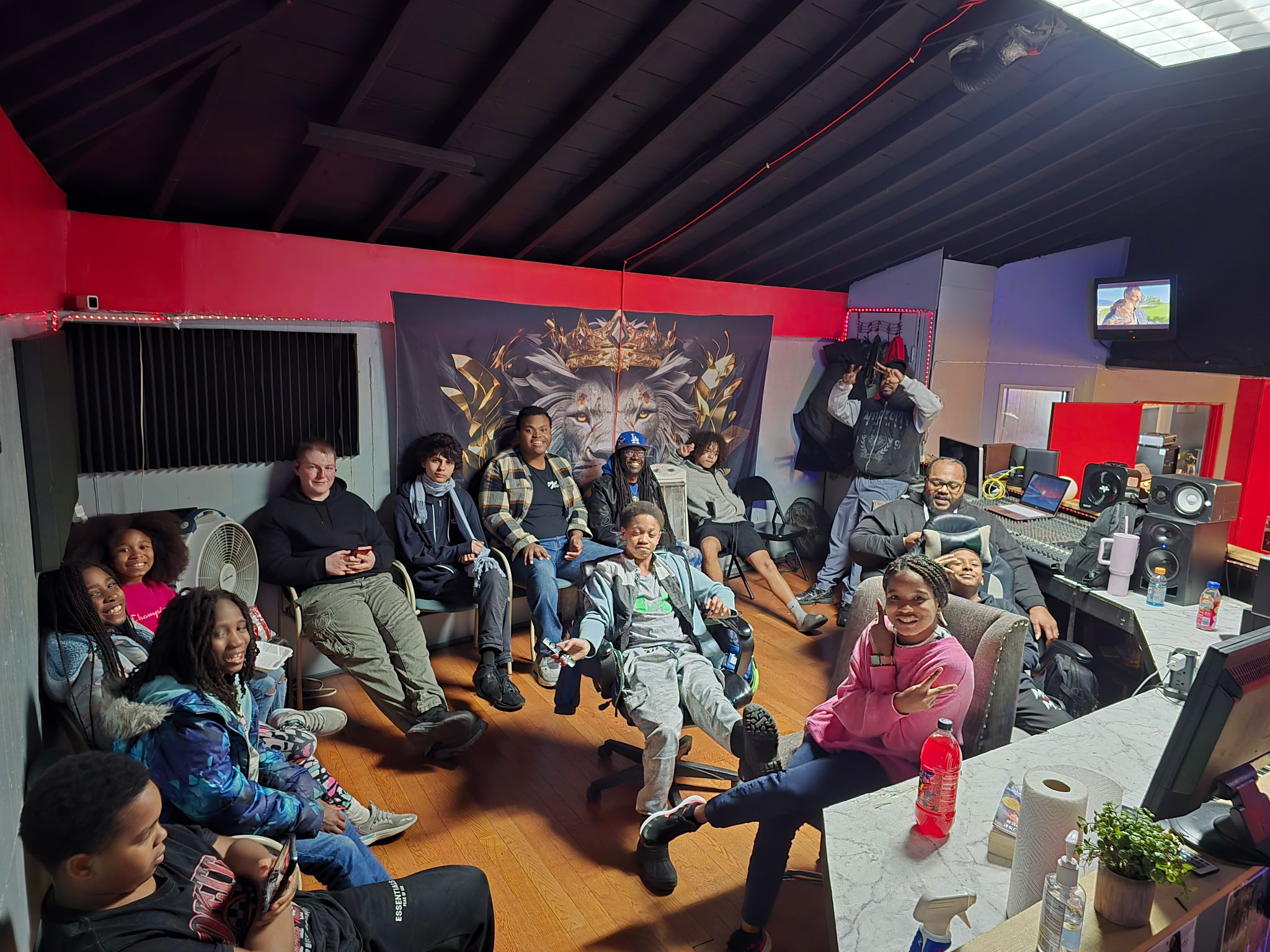 Group of people sitting and standing in a room with gaming and recording equipment, colorful walls, and a large decorative backdrop.