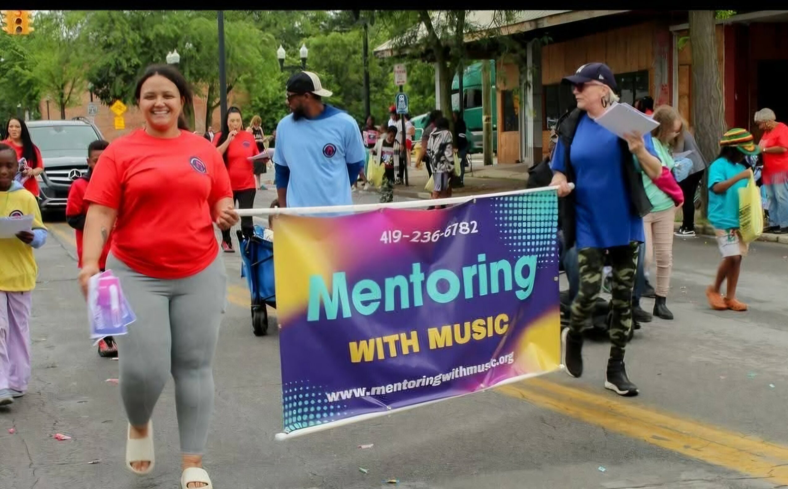 People walking on a street holding a banner that reads 'Mentoring with Music' during a community event.