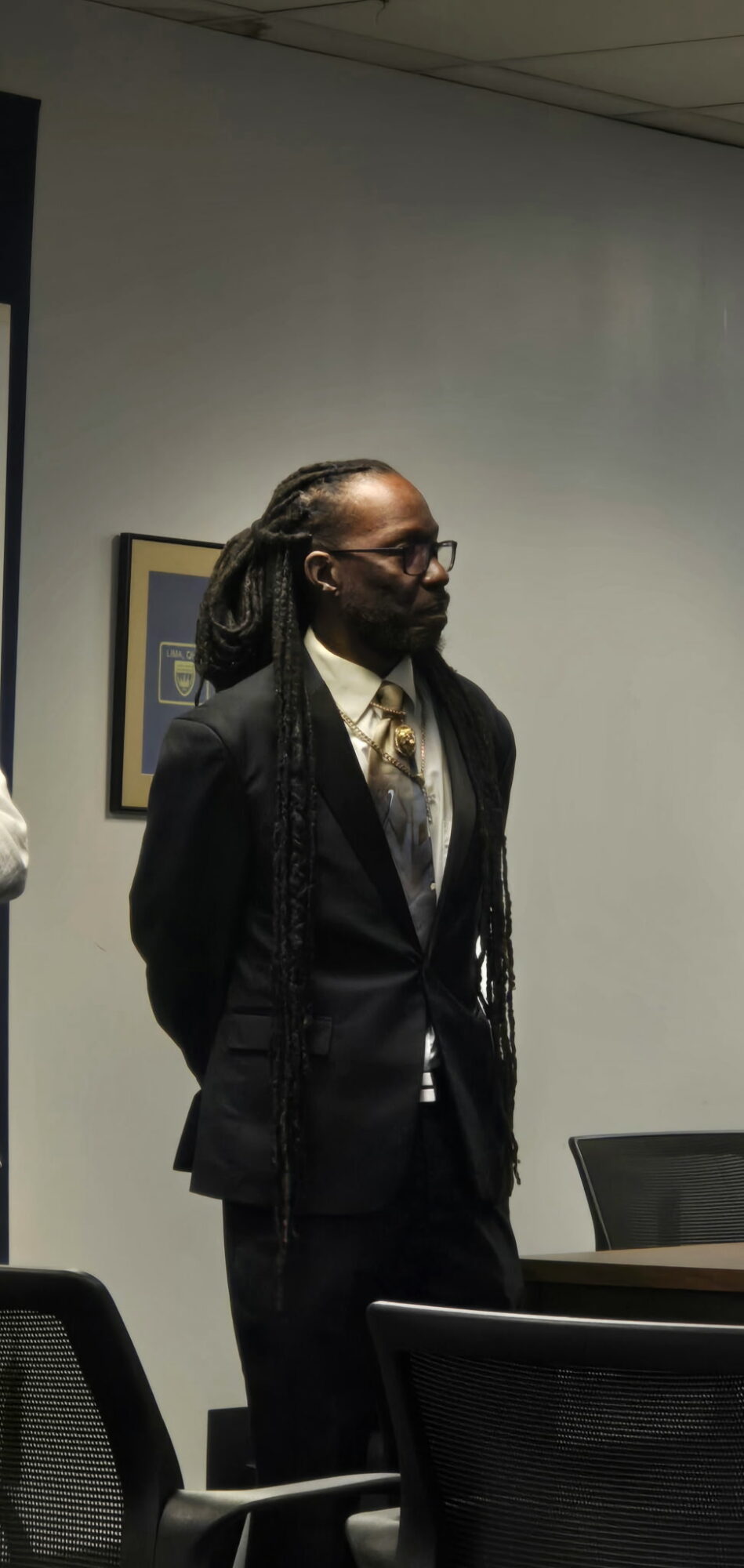 Person with glasses and braided hair stands in a room, wearing a dark suit and tie, with a framed picture on the wall.