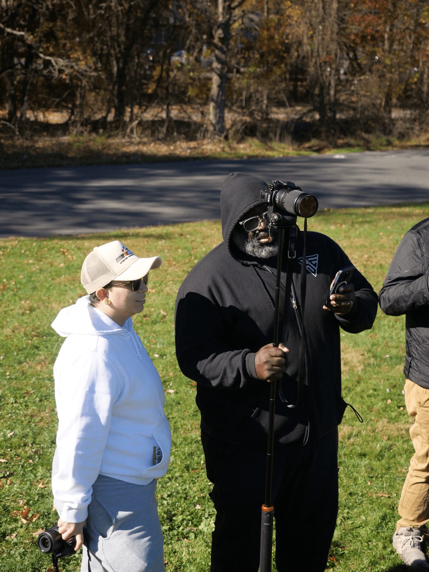 Person in black hoodie and sunglasses holding a camera, standing on grass with a child in white hoodie and cap nearby, trees in background.